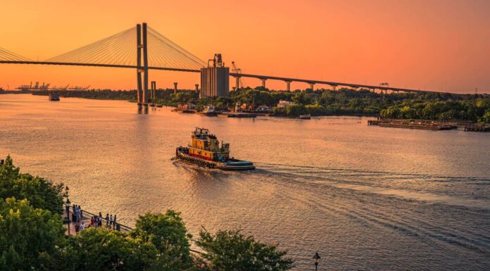 Vista artistica della sera a Savannah, in Georgia, mentre i turisti si radunano sulla riva del fiume e un rimorchiatore fa una scia dirigendosi verso il cielo dorato sotto il Talmadge Memorial Bridge.