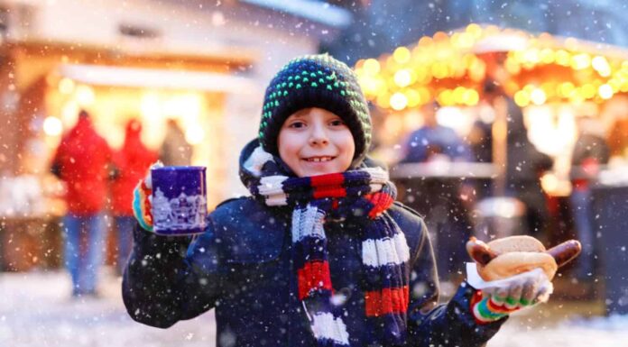 Un ragazzino carino che mangia salsiccia tedesca e beve un pugno caldo per bambini sul mercatino di Natale.  Bambino felice sul tradizionale mercato familiare in Germania.  Ragazzo che ride in abiti invernali colorati
