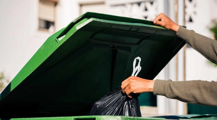 Uomo che butta fuori il sacchetto della spazzatura nero ecologico riciclabile in un grande contenitore per rifiuti verde in plastica.  Porta fuori la spazzatura