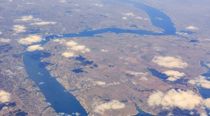 Una veduta aerea del lago Sakakawea.  Il lago è stretto e serpeggia attraverso l'inquadratura dall'inquadratura in basso a destra attraverso la parte sinistra della fotografia e poi torna a destra per circa 3/4 della salita e poi in alto verso destra ci sono alcune nuvole tra la macchina fotografica e la fotografia e il treno è troppo lontano per sembrare altro che marrone e verde.