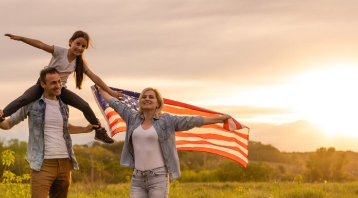 Giovani genitori con la figlia che tiene la bandiera americana in campagna al tramonto.  Celebrazione del Giorno dell'Indipendenza