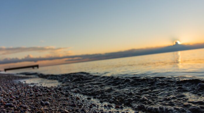 Primo piano su rocce di sabbia e piccole onde con messa a fuoco morbida sul molo di legno e sole nascente che penetra attraverso nuvole scure sopra l'orizzonte e il lago Saint-Jean, Quebec, Canada, in una calda mattina estiva