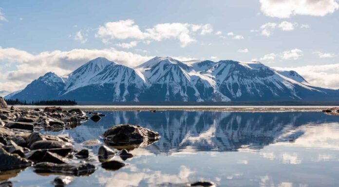Splendida vista sui Monti Atlin nella Columbia Britannica settentrionale durante la primavera in una perfetta giornata di cielo azzurro con nuvole e riflessi nell'acqua calma sottostante.  Scenico, vista per casa, ufficio art. 