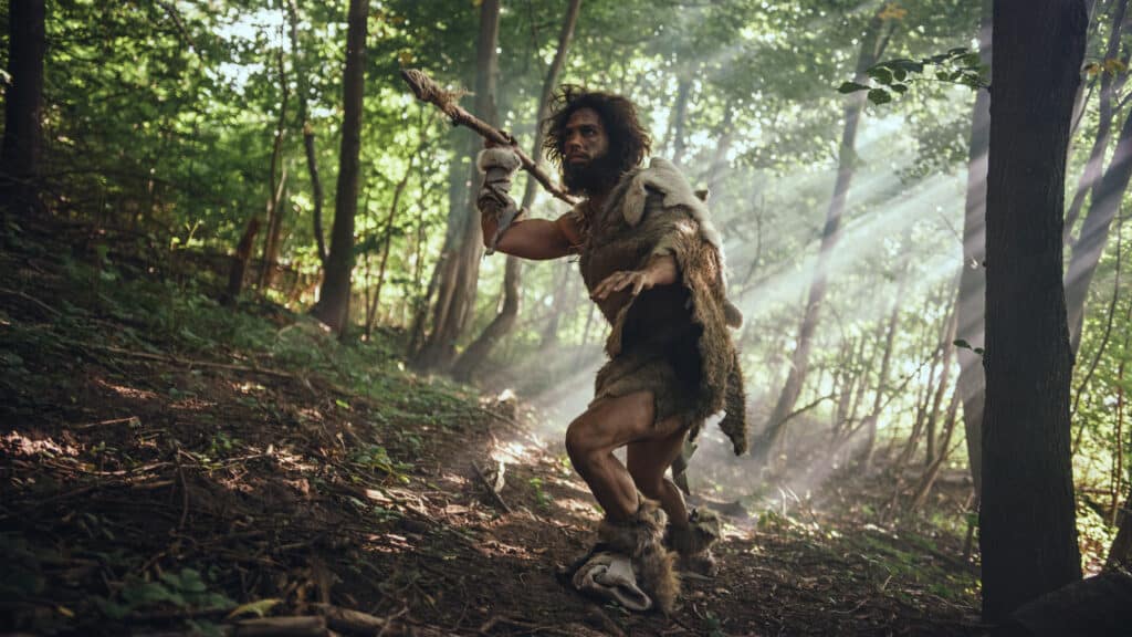 L'uomo delle caverne primordiale che indossa pelle di animale tiene una lancia con la punta di pietra si guarda intorno ed esplora la foresta preistorica a caccia di prede animali.  Neanderthal va a caccia nella giungla