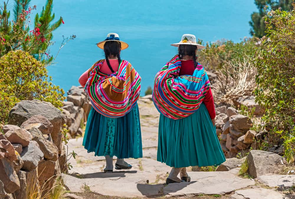 Due donne indigene Quechua in abiti tradizionali che camminano lungo il sentiero per il porto di Isla Taquile (isola di Taquile) con il lago Titicaca sullo sfondo, Perù.