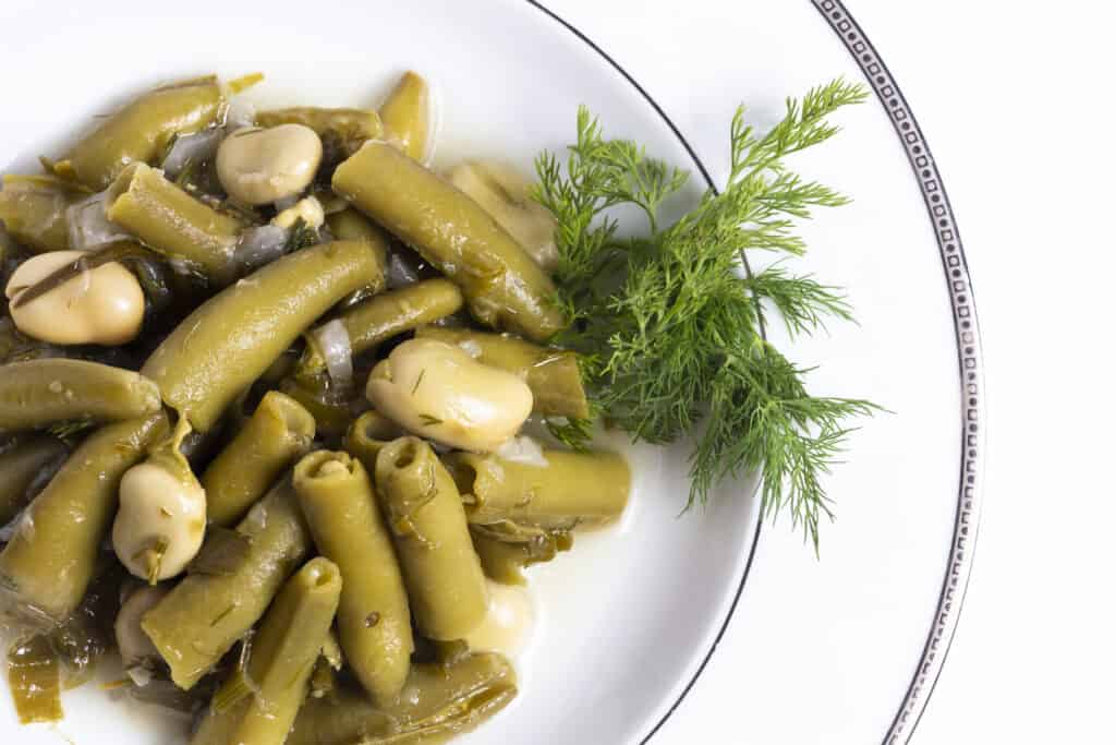 Fave colorate verde oliva cotte e patate bianche su un piatto bianco con un rametto di aneto.