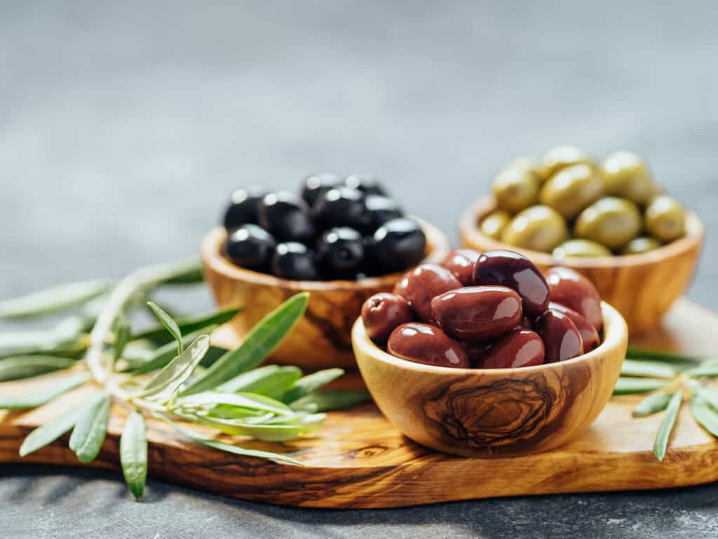 tre ciotole Woden, ciascuna di olive nere mature, kalamata e verdi su un tagliere di legno ornato di rosmarino.