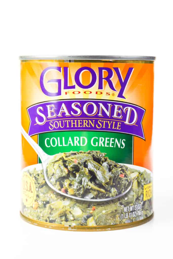 Can of Glory Foods Collard Greens condito in stile meridionale su sfondo bianco.  L'etichetta della lattina è colorata.  Il tardivo è arancione con riflessi verdi e viola.  Nella parte inferiore dell'etichetta c'è una grande ciotola di verde cotto, con una grande cucchiaiata di verdure che copre sopra il centro della ciotola.