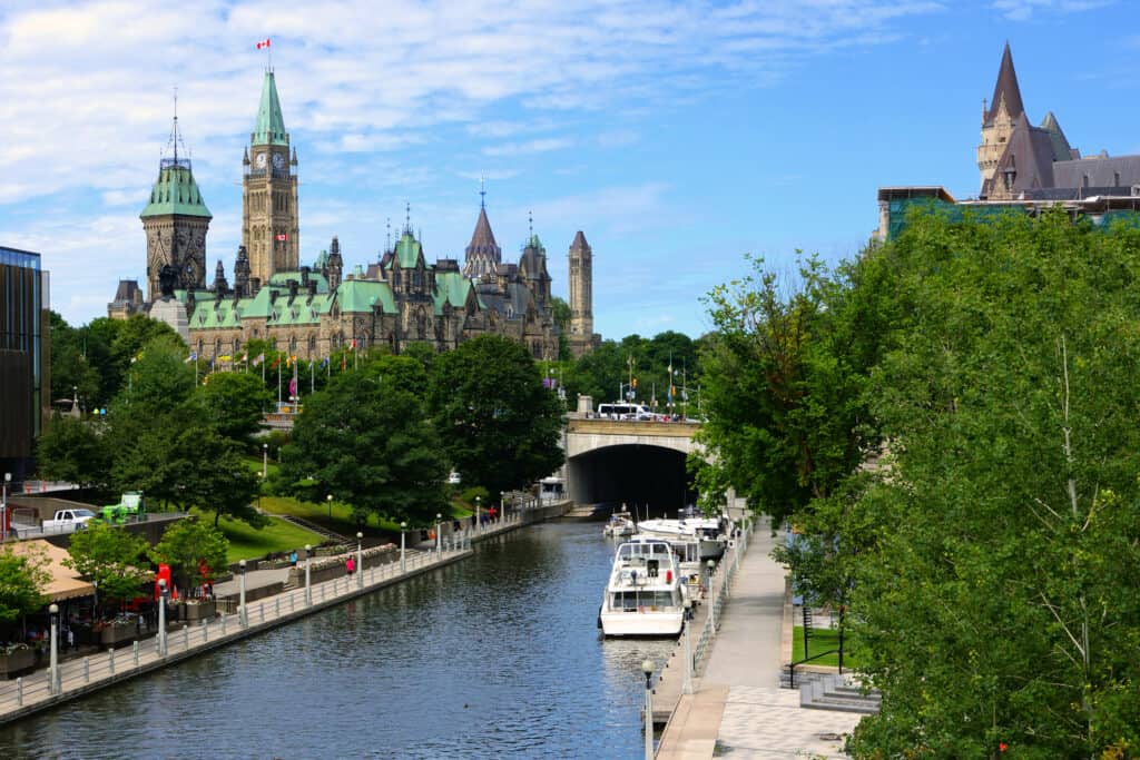 Il canale Rideau a Ottawa, in Canada in estate.  Alberi verdi fiancheggiano il canale.  due barche sono ancorate sul lato destro del canale e sullo sfondo si può vedere la collina del parlamento di Ottawa.  Un vecchio edificio con torrette e tetto color dentifricio alla menta. 