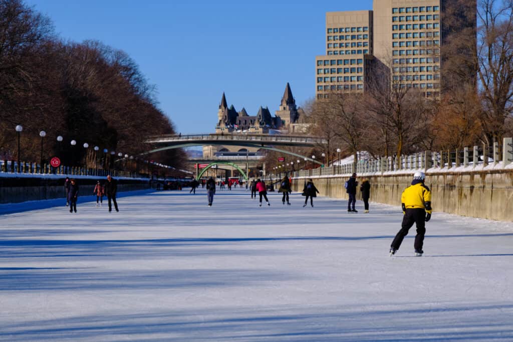 La pista di pattinaggio sul ghiaccio più grande del mondo è la Rideau Canal Skateway a Ottawa, in Canada.  Nell'inquadratura si possono vedere 10 pattinatori, anche se ce ne sono altri in lontananza.  Il pattinatore principale nell'inquadratura indossa una giacca giallo brillante e pantaloni scuri.  Paesaggio urbano di Ottawa, edificio moderno e antico sullo sfondo.  Un edificio cinque torrette visibili. 