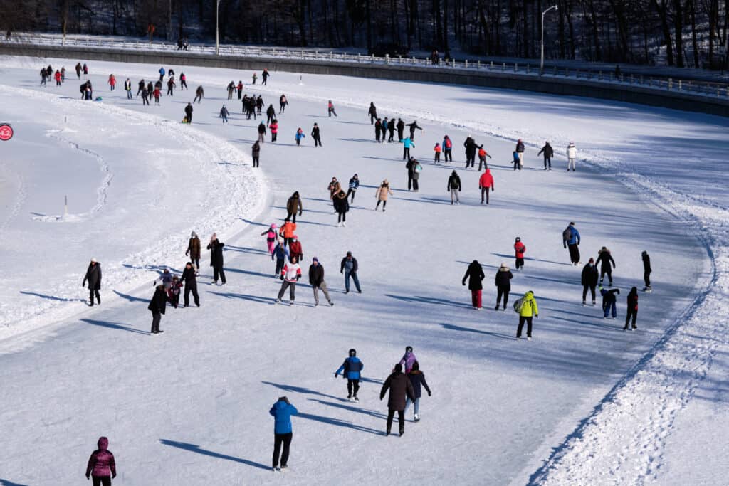 La pista di pattinaggio sul ghiaccio più grande del mondo è la Rideau Canal Skateway a Ottawa, in Canada.  Quasi 100 persone in abiti dai colori vivaci e scuri, che pattinano su un canale ghiacciato e innevato;  stanno pattinando in senso antiorario, spostandosi verso l'angolo in alto a sinistra dell'inquadratura. 