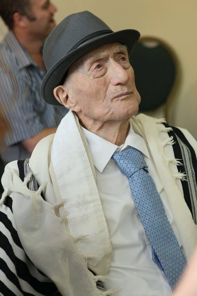 un ritratto di Yisrael Kristal, un tempo l'uomo vivente più anziano del mondo.  Kristtal ha la pelle chiara, indossa un cappello fedora grigio scuro, uno scialle da preghiera, una camicia bianca e una cravatta azzurra. 