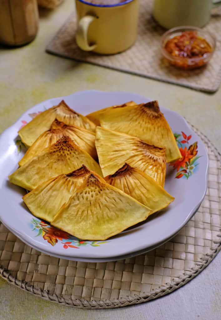 nove triangoli affettati sottilmente di pane fritto e giallastro su un piatto di porcellana con accenti floreali.  sopra una tovaglietta di canna intrecciata.  Lo sfondo è costituito da una tazza di ceramica gialla con un bordo cobalto e un piccolo piatto per condimenti in vetro trasparente che contiene una salsa rosso scuro.  Sia la tazza che il piatto di condimento sono anche su una tovaglietta intrecciata/.