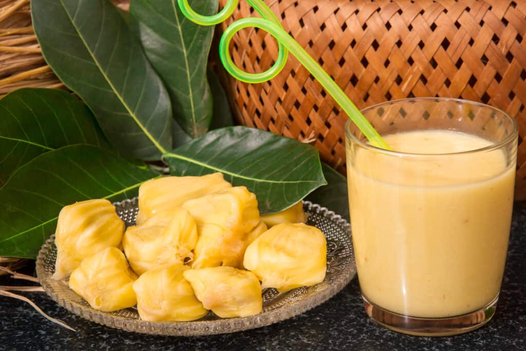 Succo di jackfruit in un bicchiere accanto a fette di jackfruit su un piatto di vetro, con presumibilmente foglie di jackfruit sopra e dietro il piatto;  sullo sfondo un cesto intrecciato