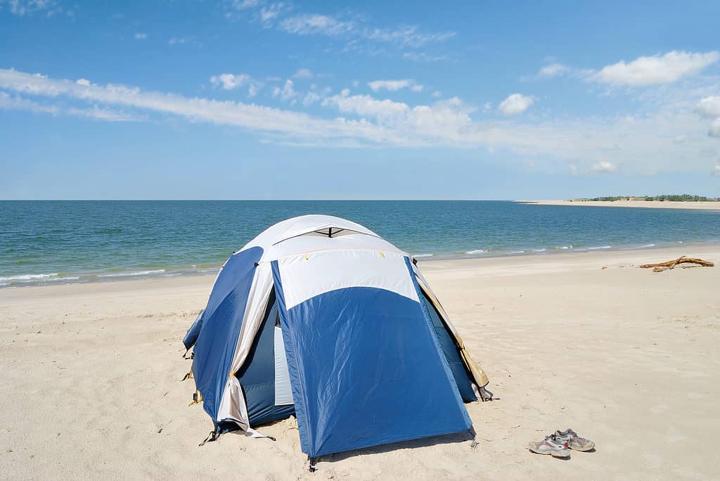 Foto di una tenda a cupola blu e argento su una spiaggia di un lago di sabbia bianca.  C'è un paio di scarpe da corsa allacciate visibili vicino alla tenda.  Il lago e un cielo azzurro con qualche nuvoletta bianca e sottile fanno da sfondo.