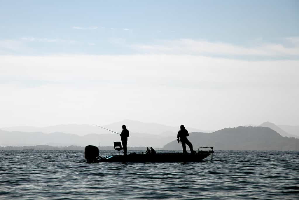 Riquadro centrale della foto: sagoma scura di due uomini in piedi che pescano in una piccola barca a motore fuoribordo su un lago.
