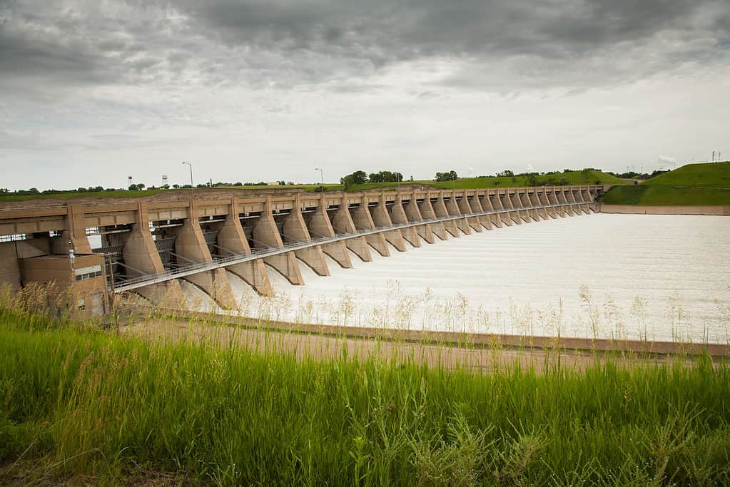 Foto di una diga su un fiume.  La diga è lunga e fatta di cemento.  C'è acqua che scorre attraverso la diga.  in primo piano sta crescendo l'erba verde alta della prateria 
