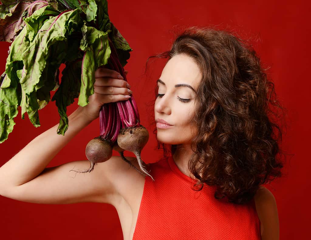 Bella giovane donna sportiva castana dalla pelle chiara in posa con foglie fresche di barbabietola verde.  Dieta.  Concetto di mangiare sano su sfondo rosso