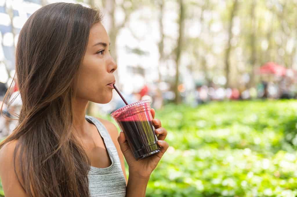 Una donna dalla carnagione olivastra con zigomi alti e lunghi capelli scuri, che indossa una t-shirt grigia senza maniche con strisce tenui, è visibile nell'inquadratura di sinistra mentre beve frullato di succo di barbabietola mentre cammina nel parco cittadino.