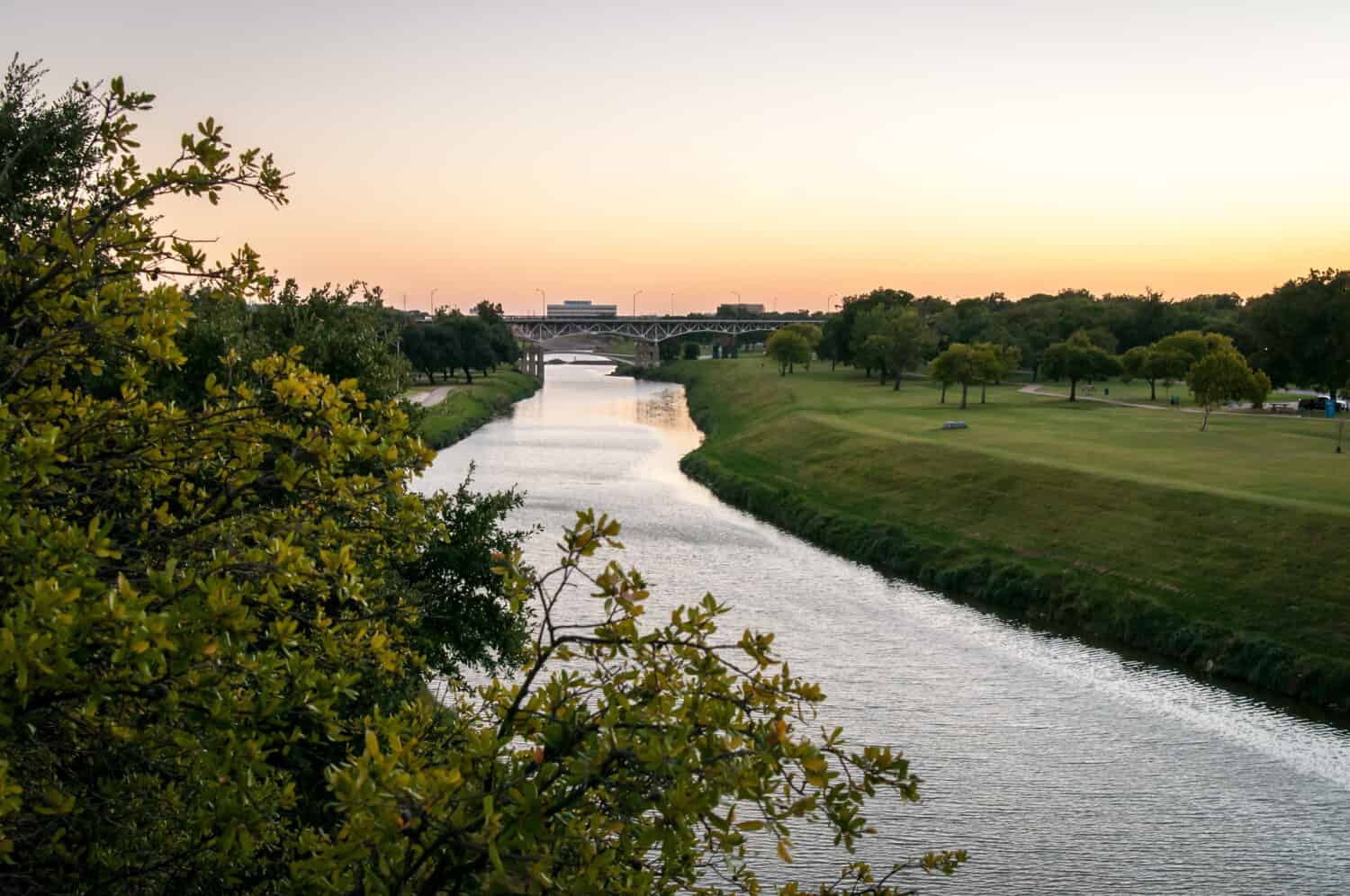 Trinità Fiume Fort Worth, Texas.  Tramonto sull'acqua, colori arancioni e dorati.  Erba verde e alberi in un parco pubblico.  Sera d'estate tranquilla e calma.  Orientamento orizzontale.
