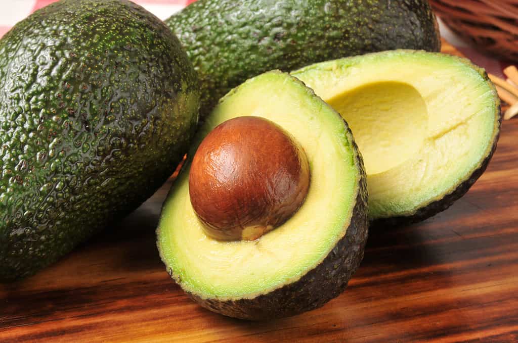 Un avocado tagliato a metà è visibile su un tagliere di legno.  La fossa è ancora nella metà più vicina alla lente.  Due avocado interi e non tagliati sono visibili nella cornice dietro l'avocado affettato.