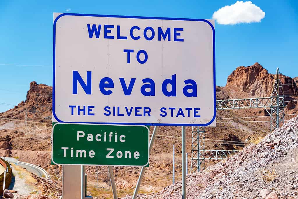 Benvenuti al cartello del Nevada al confine tra Nevada e Arizona vicino alla diga di Hoover, il cartello è bianco con scritte blu.  Un piccolo segno verde nella cornice in basso a sinistra dice "Fuso orario del Pacifico" in lettere bianche. 