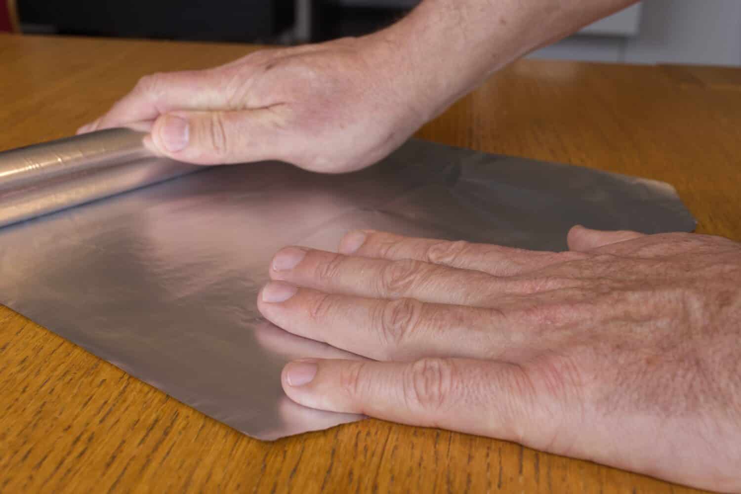 Le mani degli uomini rotolano via il foglio di alluminio per uso domestico su una superficie di legno.