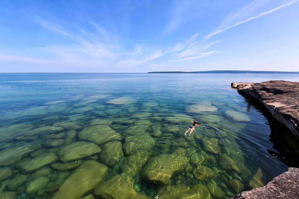 Una ragazza nuota nel Lago Superiore nella penisola superiore del Lago Michigan.  Le rocce sono visibili attraverso il vetro come acque incontaminate.  Nella foto Rocks National Lakeshore è in lontananza.