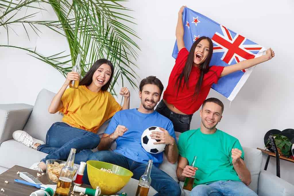 Gruppo di amici appassionati di sport che guardano la partita in camicie colorate ragazza con bandiera della nuova zelanda