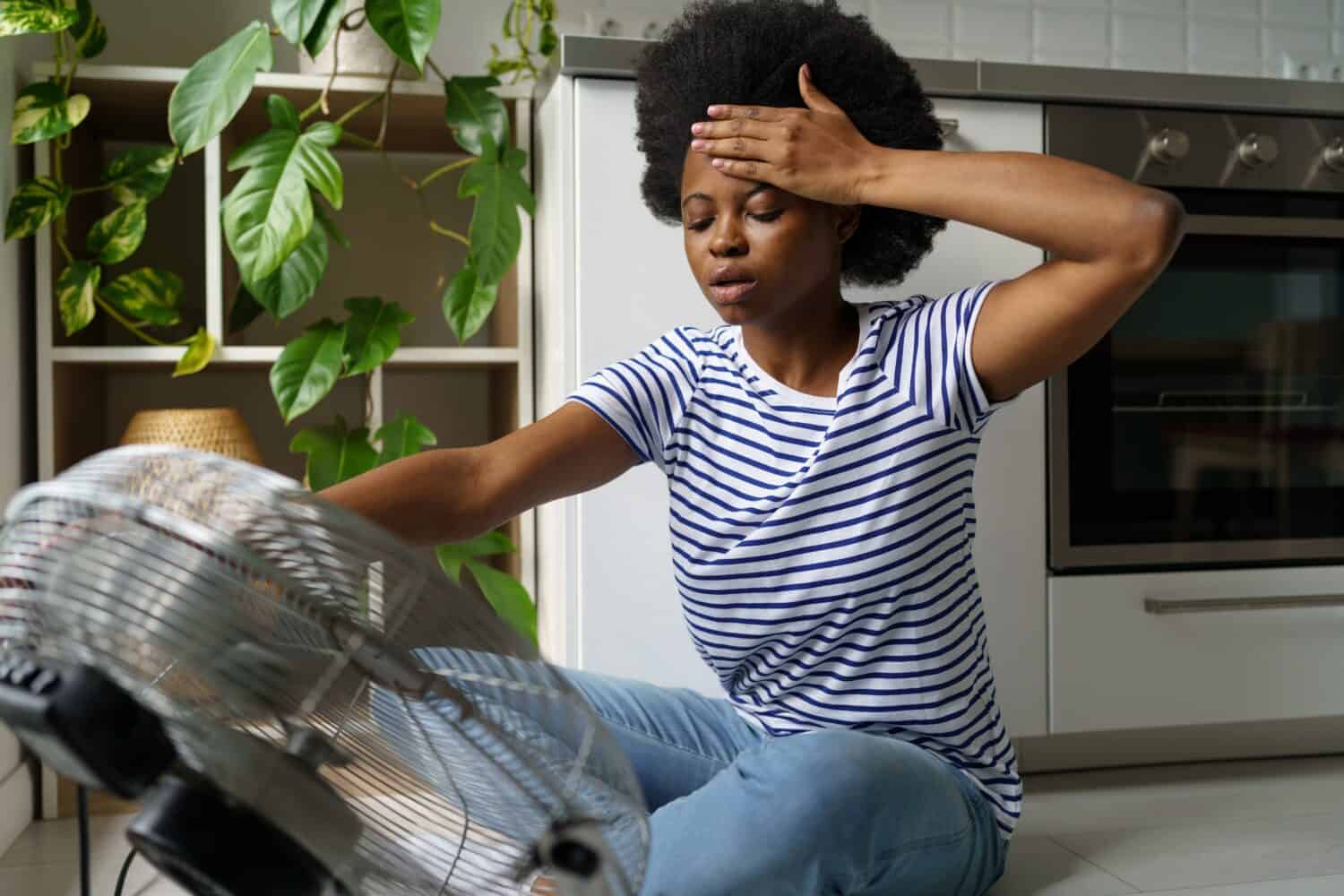 Donna africana infelice che tocca la fronte con vampate di calore usando il ventilatore elettrico per rinfrescarsi, donna nera surriscaldata seduta davanti al refrigeratore d'aria a casa, sopravvissuta all'ondata di caldo senza aria condizionata