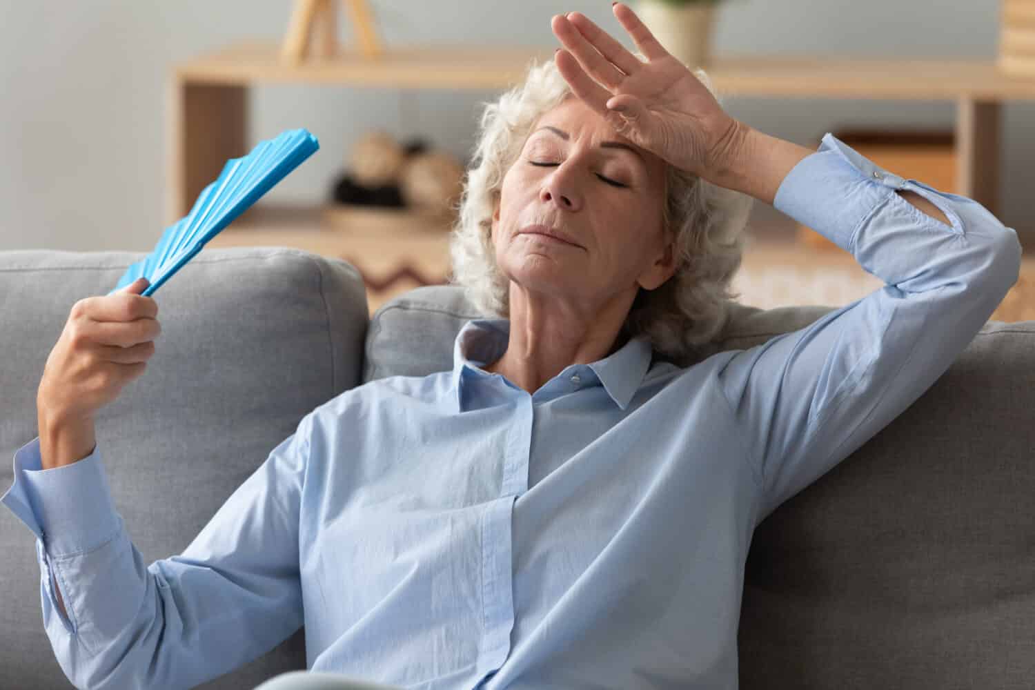 La donna anziana stanca infastidita che tiene il ventilatore ondeggiante si sente caldo seduto sul divano di casa senza condizionatore d'aria, surriscaldata esausta vecchia nonna anziana che suda soffre di problema ormonale concetto di calore estivo