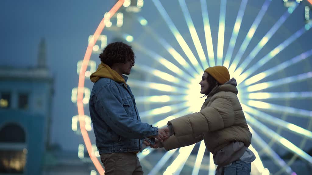 Donna allegra che flirta con il ragazzo all'appuntamento romantico all'aperto.  Coppia d'amore che si diverte contro la ruota panoramica illuminata nel centro della città.  Uomo affettuoso che abbraccia la ragazza sulla strada urbana di notte.