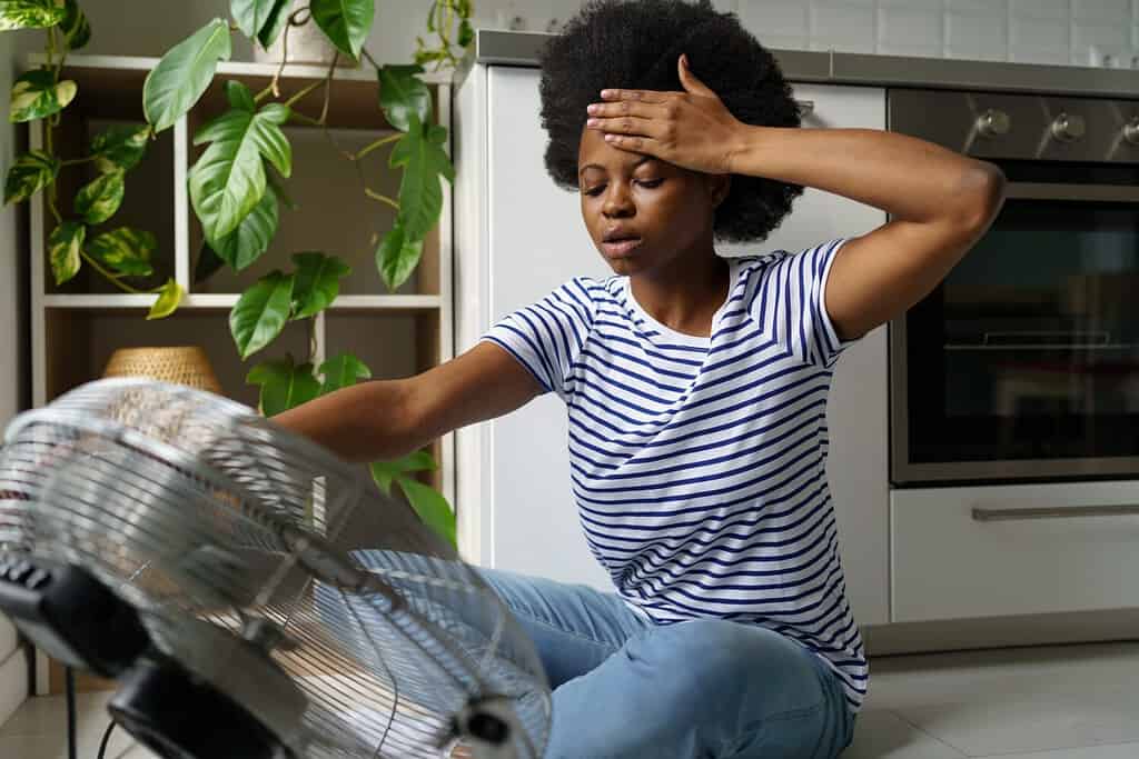 Donna africana infelice che tocca la fronte con vampate di calore usando un ventilatore elettrico per rinfrescarsi, donna nera surriscaldata seduta davanti al frigorifero dell'aria a casa, sopravvissuta all'ondata di caldo senza aria condizionata