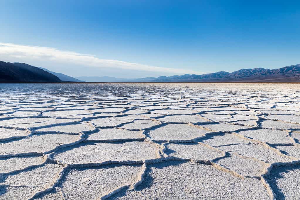 Alba sul bacino di Badwater, Death Valley, California.  Sprazzo di sole sulle montagne lontane;  il fondo della vasca è ricoperto da depositi di sale bianco;  formazioni cristalline serpeggianti formano forme esagonali in lontananza.