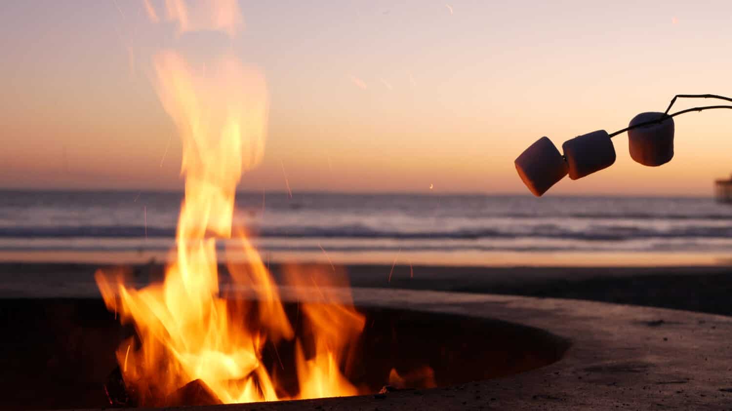 Campfire pit da Oceanside Pier, California USA.  Fuoco da campo sulla spiaggia dell'oceano, fiamma del falò in un anello di cemento per barbecue, onde di acqua di mare.  Riscaldare, arrostire o tostare marshmallow su bastoncino.  Romantico cielo al tramonto