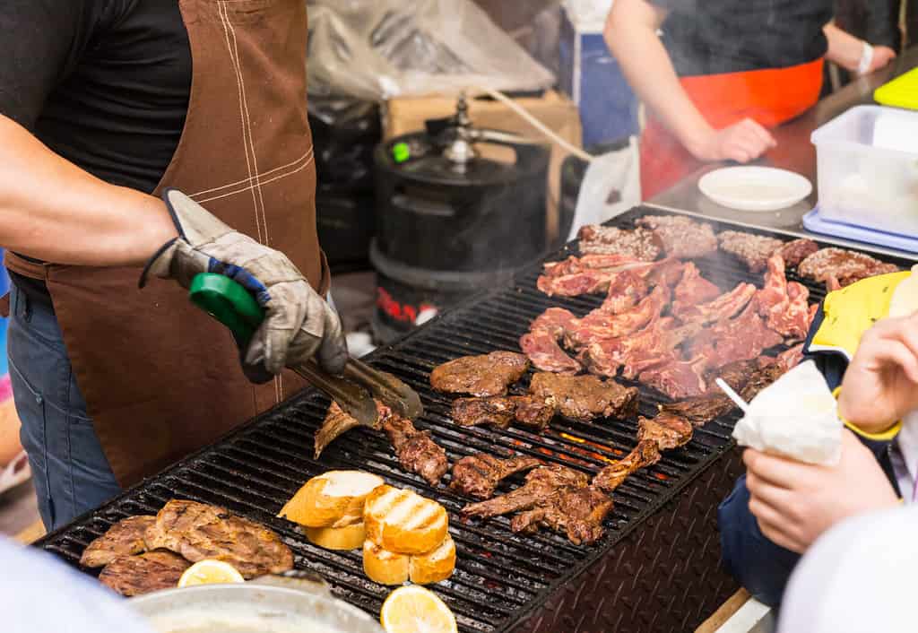 Persona che griglia carne in occasione di un evento di catering con una vista ravvicinata delle mani dello chef e della cottura della carne alla griglia sopra un fuoco