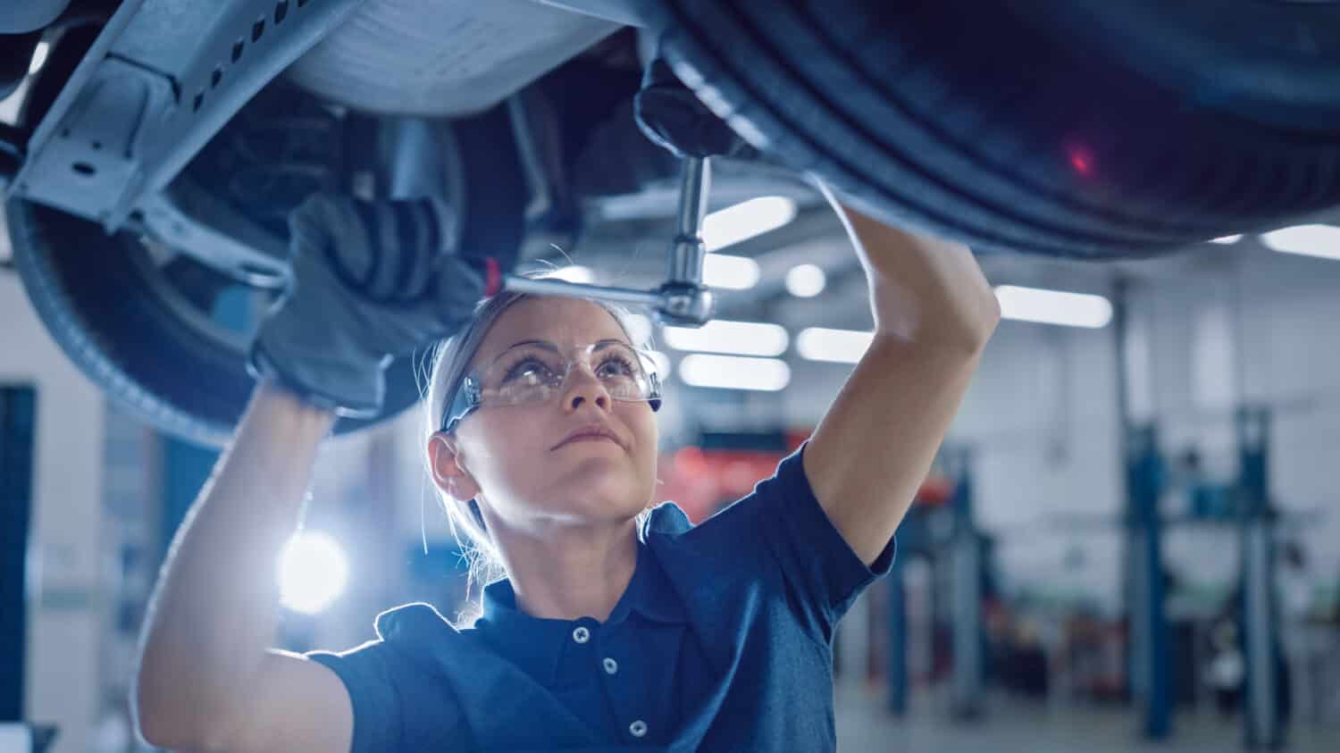 Ritratto di un meccanico femminile che lavora sotto il veicolo in un servizio di auto.  Empowering Woman Indossa guanti e utilizza un cricchetto sotto l'auto.  Officina pulita moderna.