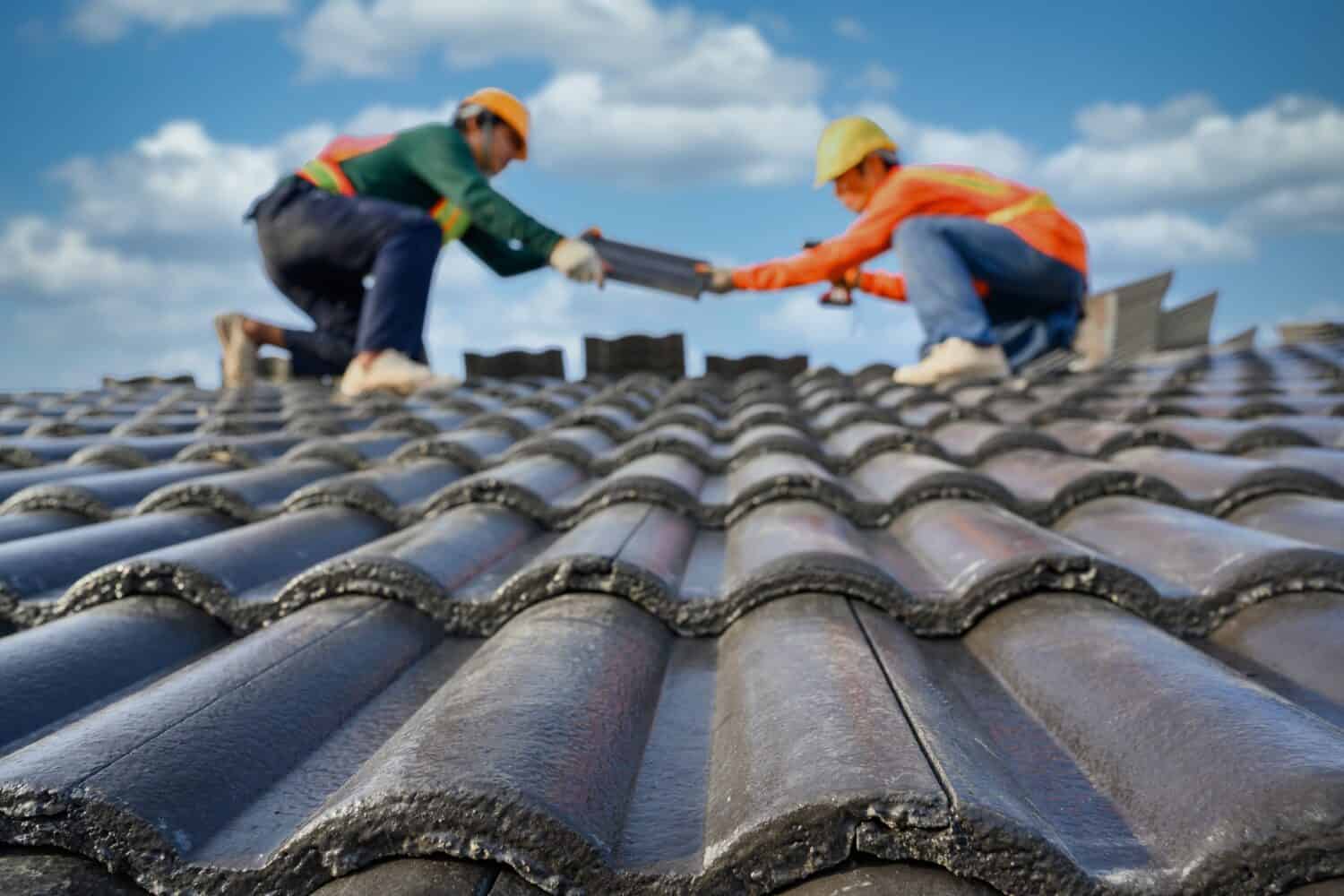 Seleziona il focus sulle tegole.  Sfondo sfocato di un roofer che lavora sul tetto di una casa Utilizzare un trapano per forare le viti per fissare le tegole di cemento.