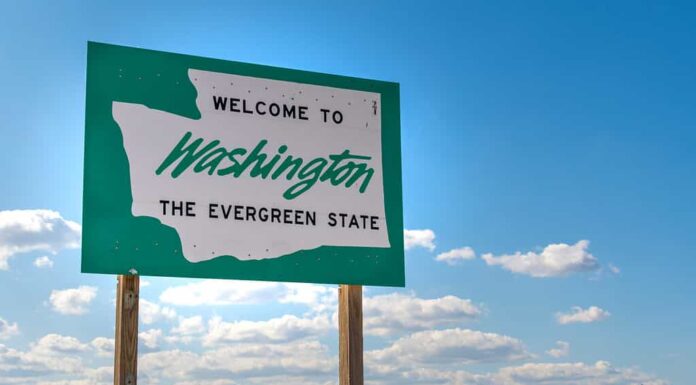 Un cartello stradale con il benvenuto nello Stato di Washington, lo stato sempreverde scritto su di esso con un ambiente rurale e un cielo blu dietro.