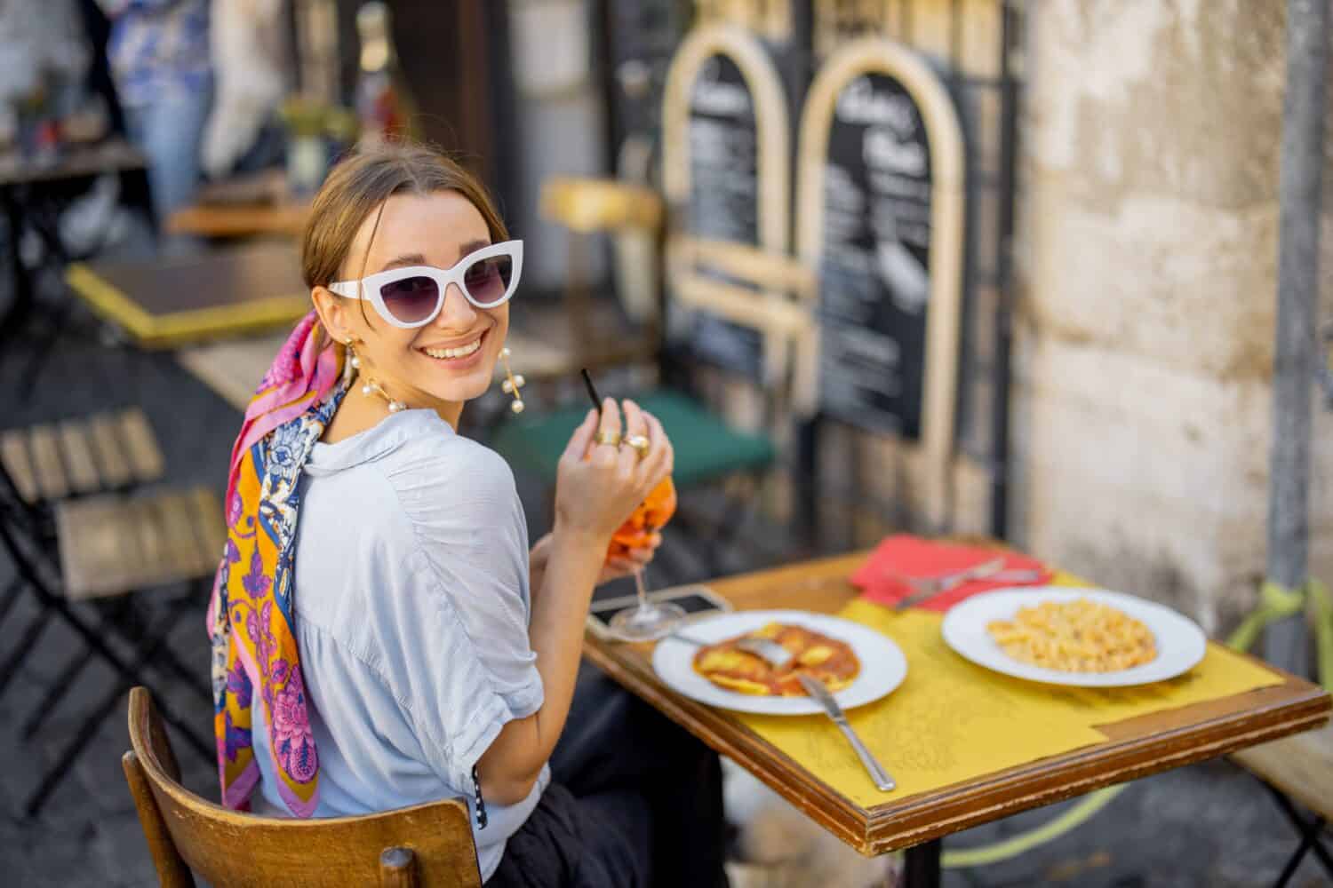 Donna che mangia pasta italiana e beve vino al ristorante sulla strada a Roma.  Concetto di gastronomia e viaggi italiani.  Donna alla moda con occhiali da sole e scialle di capelli colorati