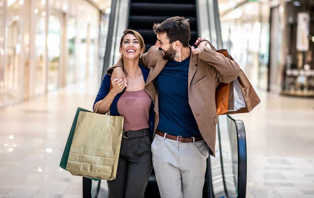 Giovani coppie felici attraenti che abbracciano, sorridono e tengono le borse della spesa mentre camminano nel centro commerciale.