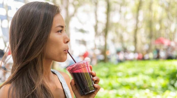 Una donna dalla carnagione olivastra con zigomi alti e lunghi capelli scuri, che indossa una t-shirt grigia senza maniche con strisce tenui, è visibile nell'inquadratura di sinistra mentre beve frullato di succo di barbabietola mentre cammina nel parco cittadino.