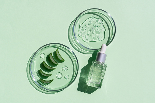 Disposizione piatta di siero cosmetico e vetreria da laboratorio con foglie fresche di aloe vera su sfondo verde brillante.