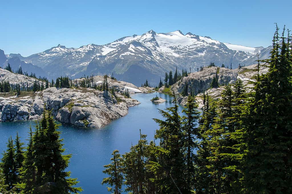 Pittoresco scintillante lago Robin circondato da granito e alberi con il Monte Daniel innevato che torreggia sullo sfondo sotto un cielo blu senza nuvole nel deserto dei laghi alpini, nello Stato di Washington, USA