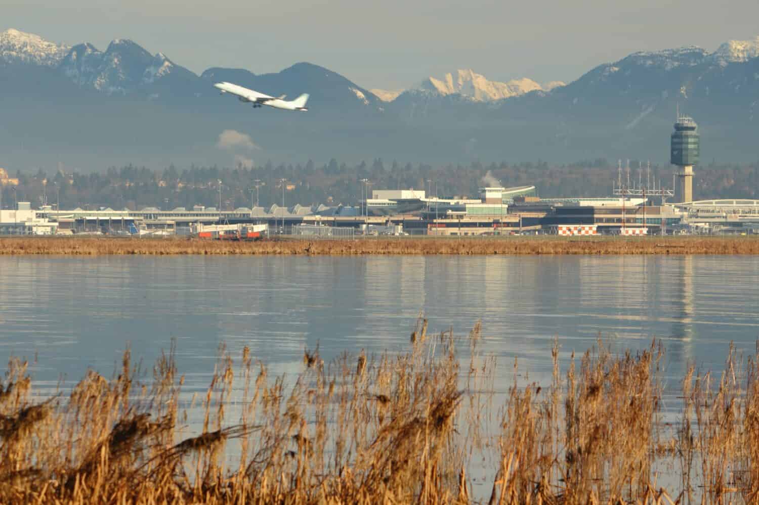 Un aereo in decollo all'Aeroporto Internazionale di Vancouver, YVR, con le Coast Mountains sullo sfondo e il fiume Fraser in primo piano.  Columbia Britannica, Canada.