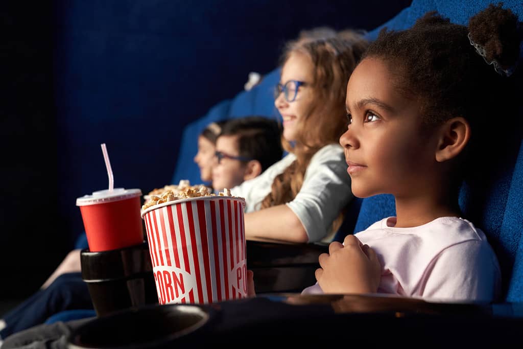 Bella ragazza africana con un'acconciatura divertente che guarda film emozionati al cinema.  Meraviglioso bambino seduto con gli amici, mangiando popcorn e sorridendo.  Concetto di intrattenimento e divertimento.