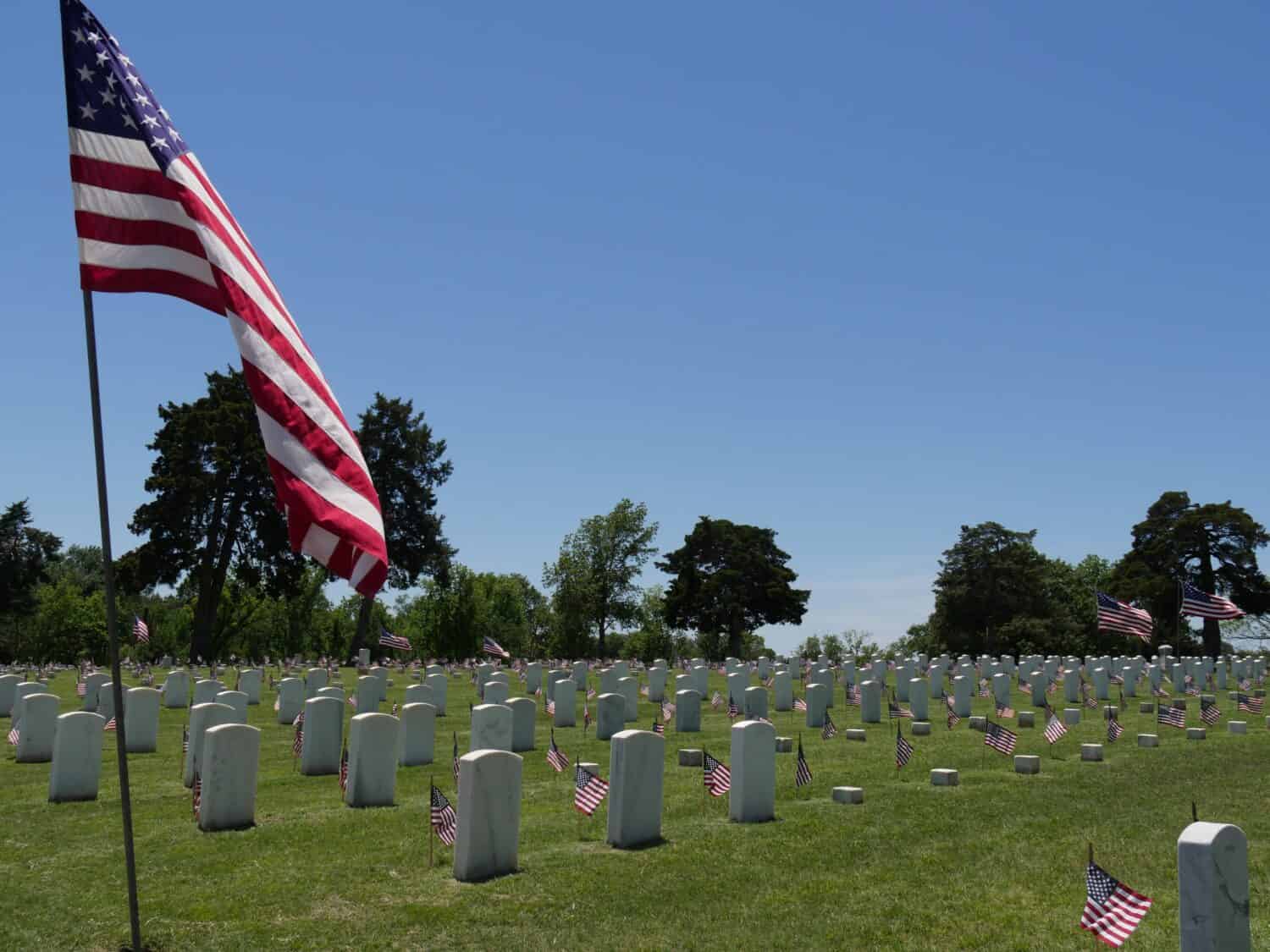 Bandiere degli Stati Uniti al cimitero commemorativo dei veteraniUna grande bandiera degli Stati Uniti a metà asta e piccole bandiere degli Stati Uniti sventolano nel vento accanto alle lapidi del cimitero nazionale di Fort Gibson a Muskogee, Oklahoma