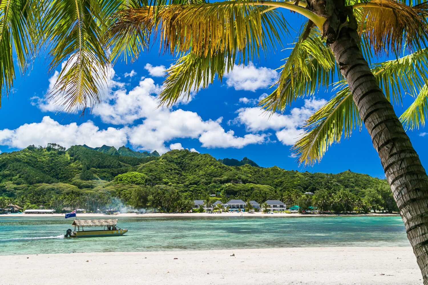 Baia tropicale, destinazione paradisiaca delle Isole Cook.  Costa di Rarotonga con palme durante una giornata di sole.  Una barca con la bandiera delle Isole Cook sul mare.  Cielo azzurro con nuvole.  Parzialmente nuvoloso.