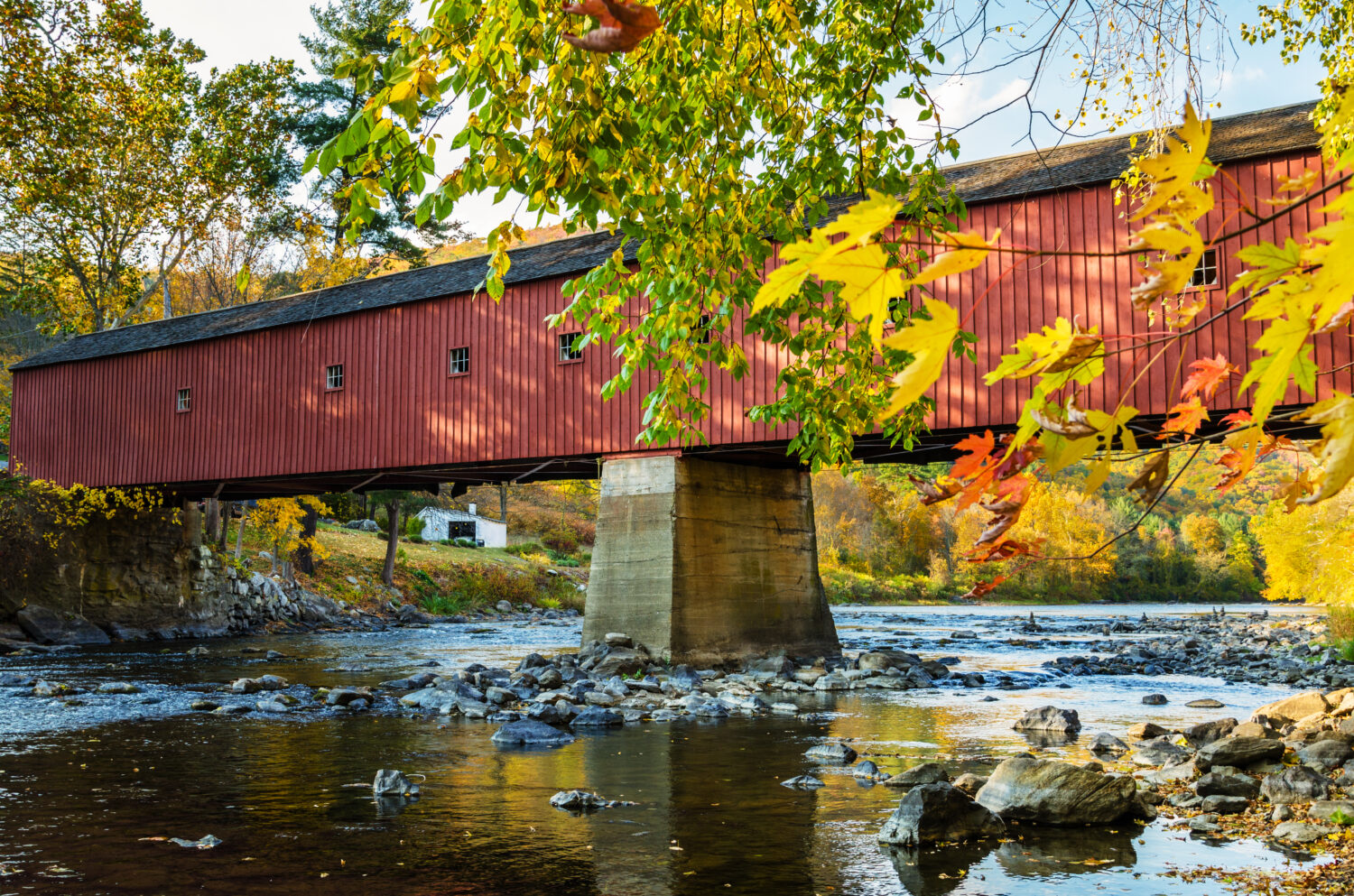 Tradizionale ponte coperto rosso che attraversa un fiume con rami di alberi in primo piano in una soleggiata giornata autunnale.  Cornovaglia occidentale, CT.