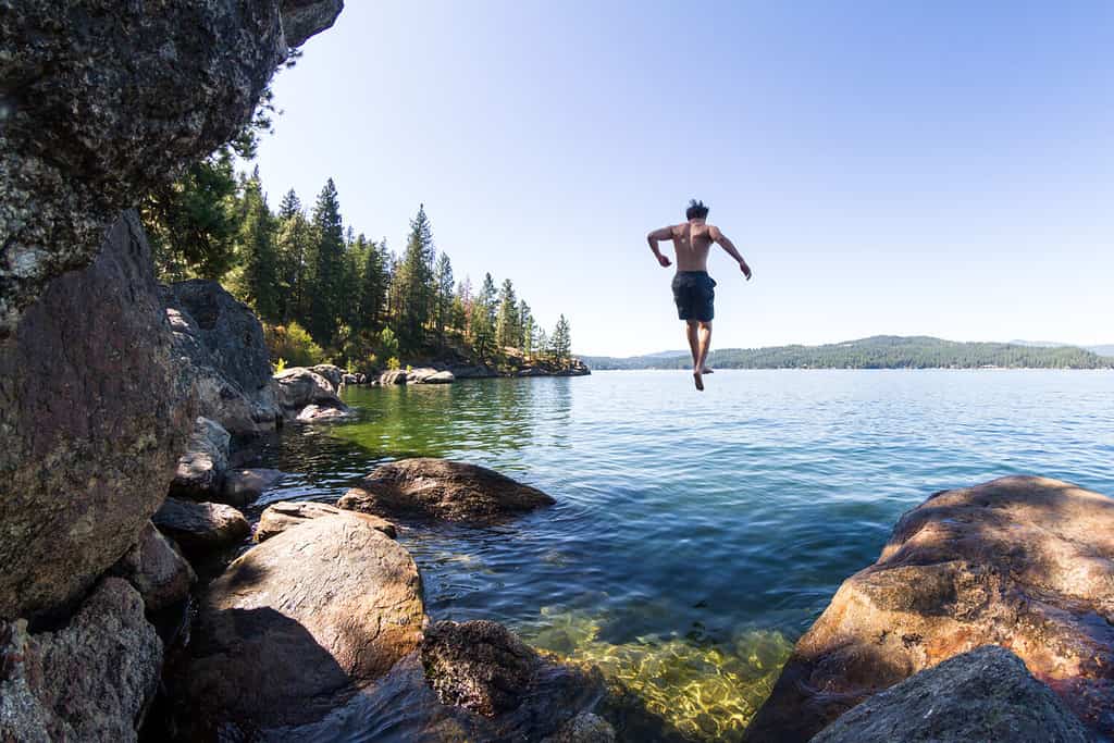 Giovane uomo che salta da una scogliera rocciosa a Tubs Hill nel lago Coeur d'Alene nell'Idaho settentrionale